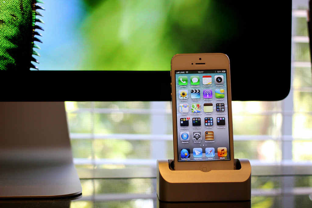 An iPhone 5 standing in an Elevation Dock in front of an iMac, home screen showing